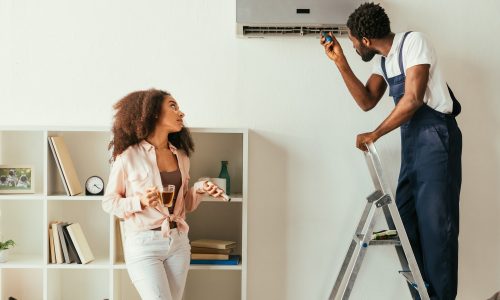 african american repairman fixing air conditioner near pretty african american woman holding remote